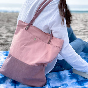 Tote bag shown in light pink color. Held by a girl with brown hair at the beach